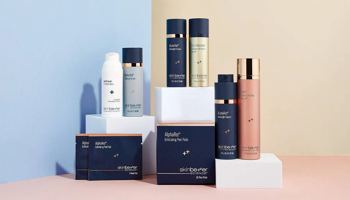 A selection of skincare products from Skinbetter Science displayed on geometric stands against a two-tone background. The arrangement includes bottles and boxes in navy and white packaging with gold accents.