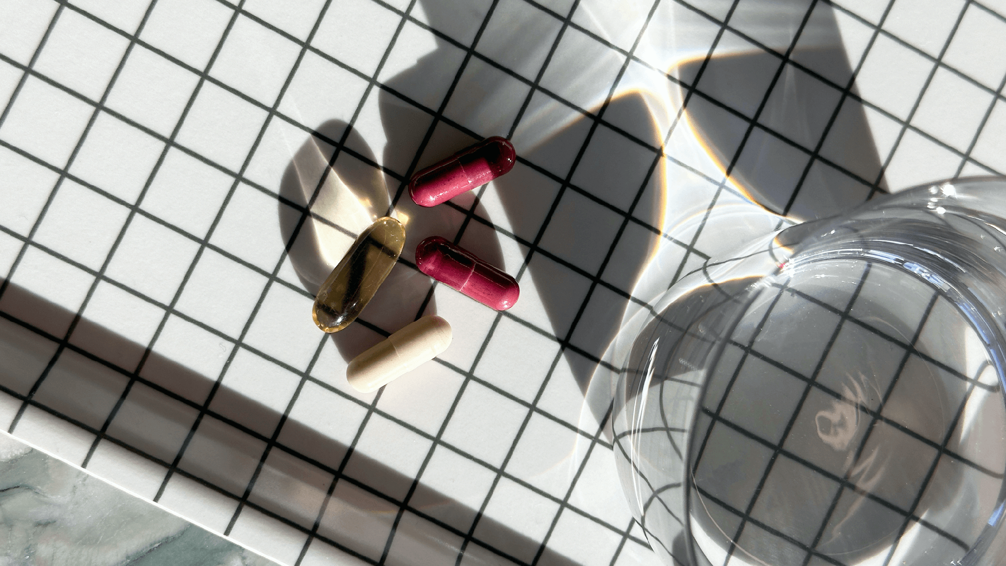 Various pills on a tiled surface with grid lines and a glass with sunlight flare and shadow.