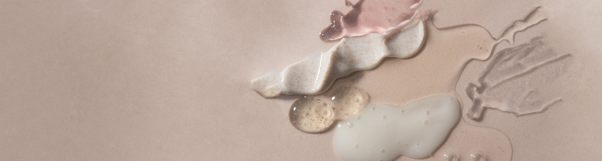 Various textures of skincare products spread on a soft pink background, showcasing creamy, gel-like, and slightly sparkly consistencies in neutral tones.