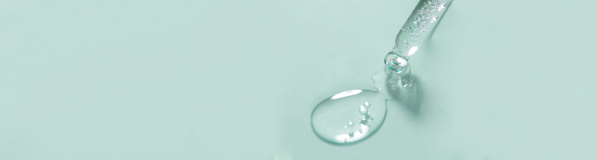 Close-up of a clear dropper releasing a droplet of liquid onto a smooth, pastel green surface. The liquid is transparent and glossy, capturing light reflections. The background is minimal, highlighting the purity and clarity of the liquid.