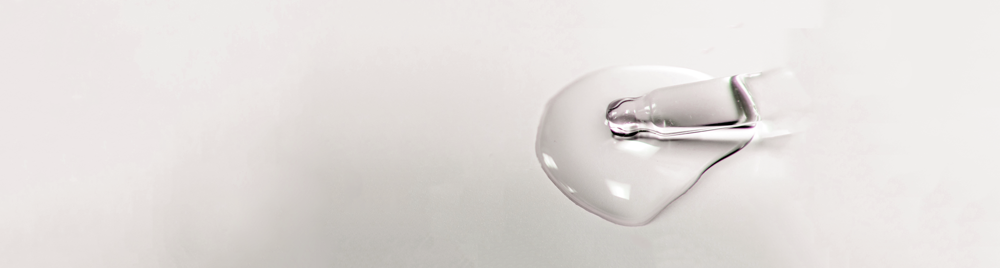 A droplet of clear liquid coming from a clear serum bottle dropper sits on a smooth, reflective surface. The background is a soft white, emphasizing a transparency and fluidity of the droplet.