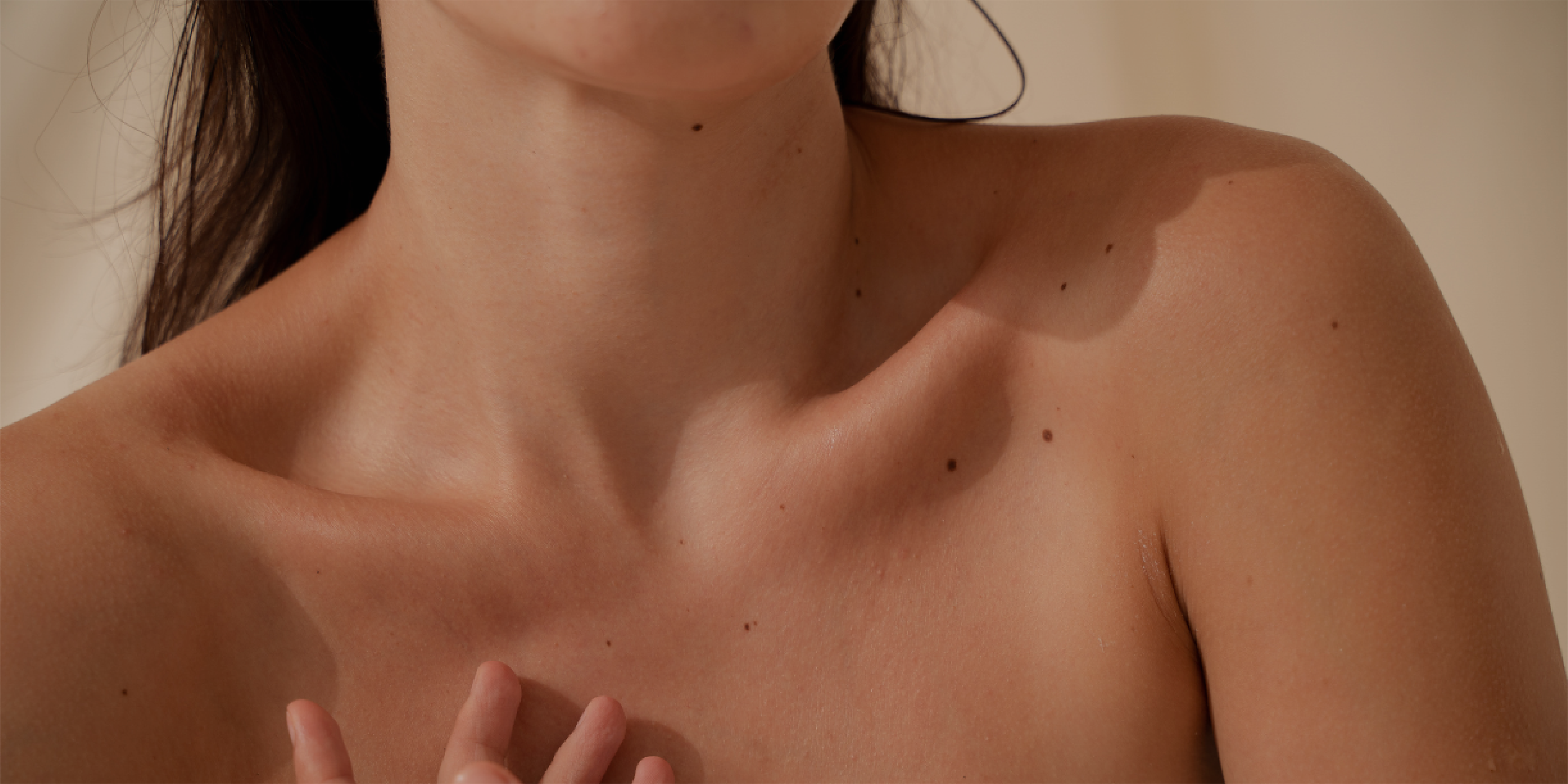 Close-up of a persons upper chest and neck, showcasing the collarbone and glowing tanned the skin. The background is softly blurred, focusing on the skins texture and natural features. The person has dark hair.