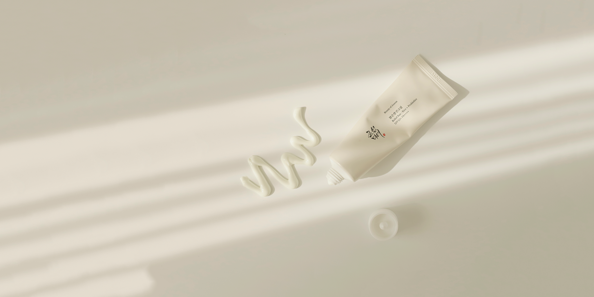 A white tube of cream from Beauty of Joseon with its cap off lies on a sunlit surface. Cream is artistically squeezed out in a wavy line next to the tube.