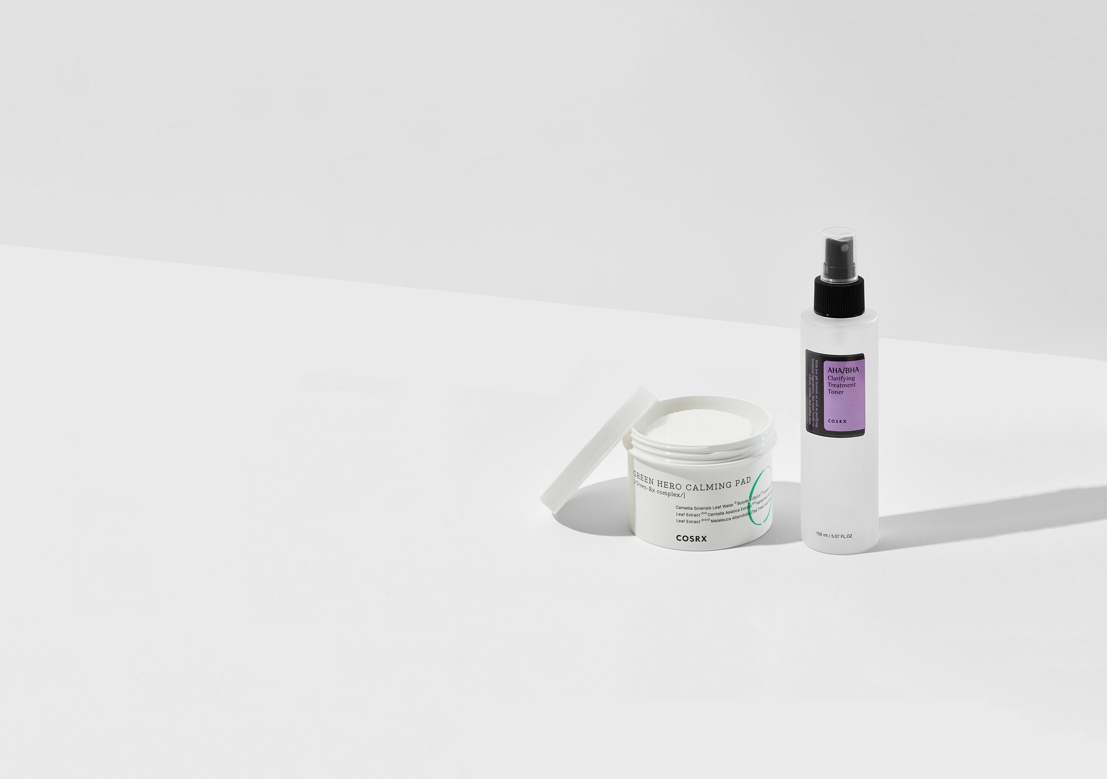 Minimalist image of a COSRX skincare set: a white jar labeled One Step Green Hero Calming Pads with the lid slightly open, and a spray bottle labeled Hyaluronic Acid Hydra Power Essence standing upright on a white surface.