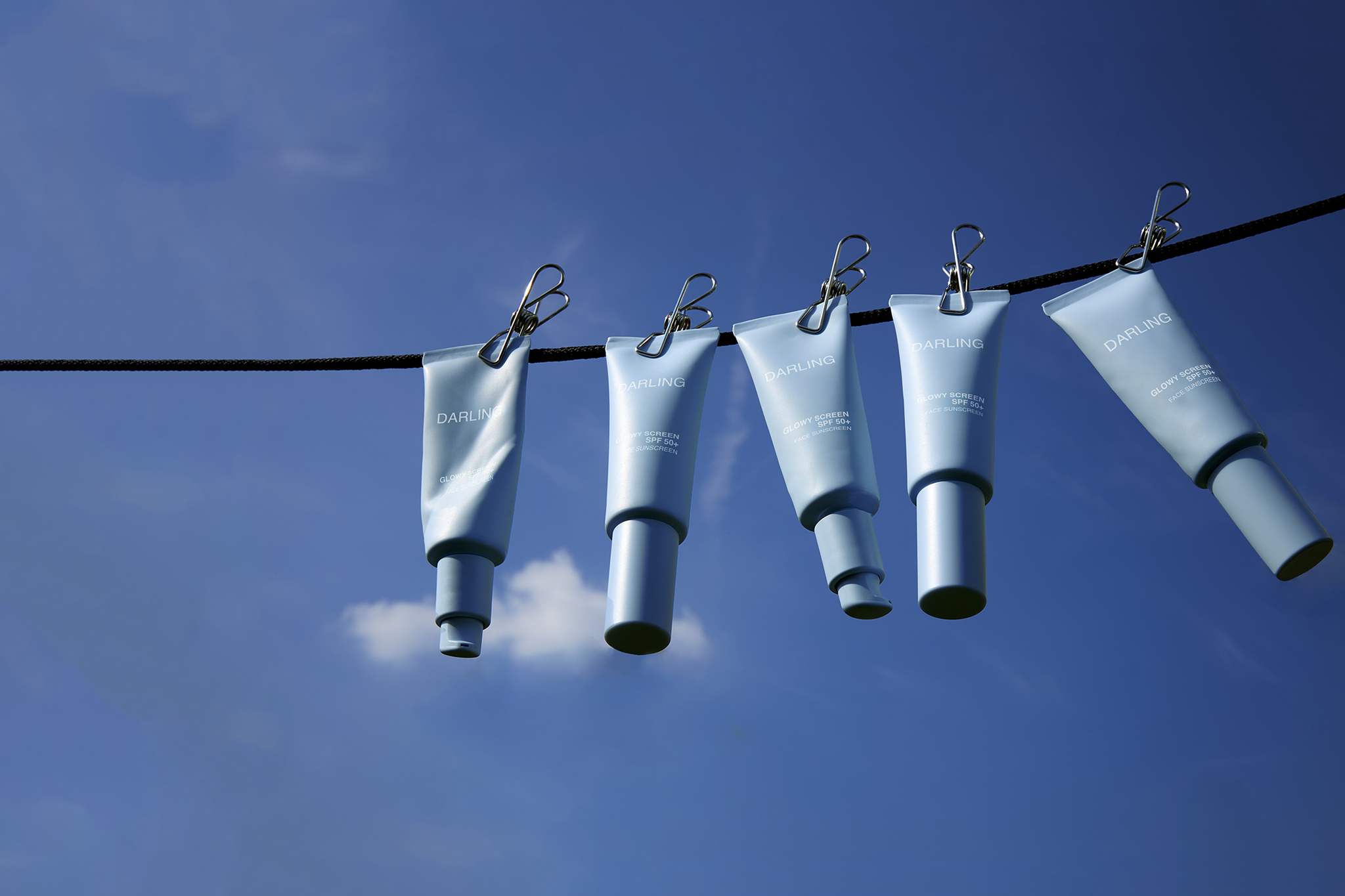 Five blue tubes from Darling with silver clips hang from a black wire against a clear blue sky. Each tube is labeled Darling and arranged vertically, creating a unique visual against the minimal background.