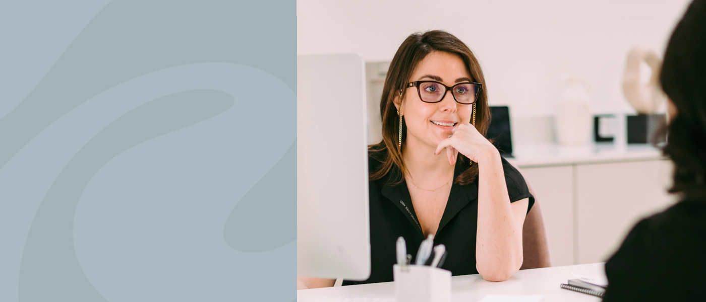 An office scenario with an individual facing another at a minimalistic white desk. Focus on smiling woman.