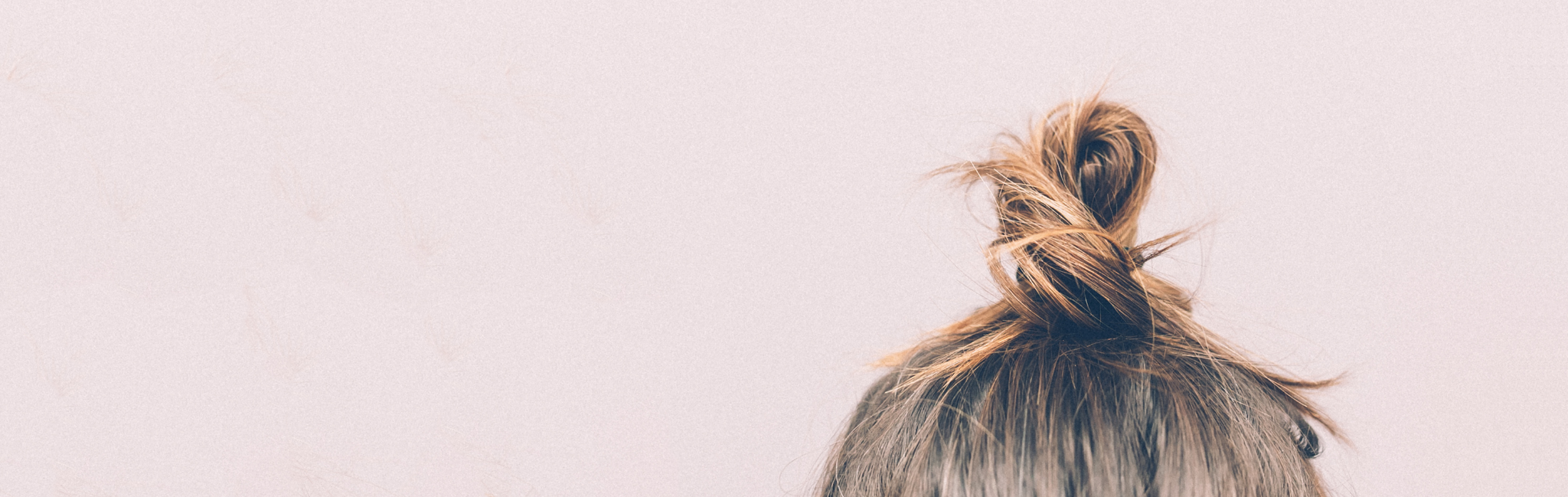 Close-up image of the back of a head with light brown hair tied up into a bun on top of the head. The background in light mauve.