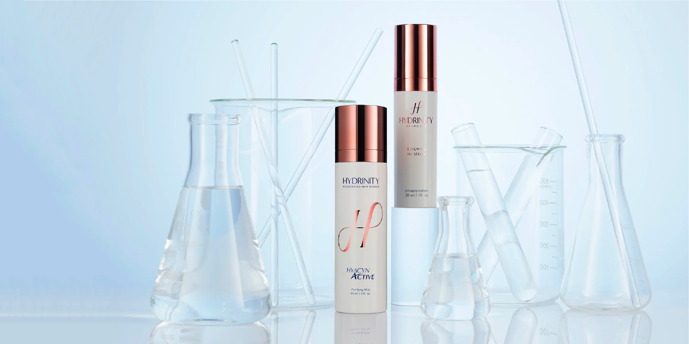 Two cylindrical skincare product bottles from Hydrinity with copper-colored caps are placed among various clear laboratory glassware containing water, set against a light blue background.