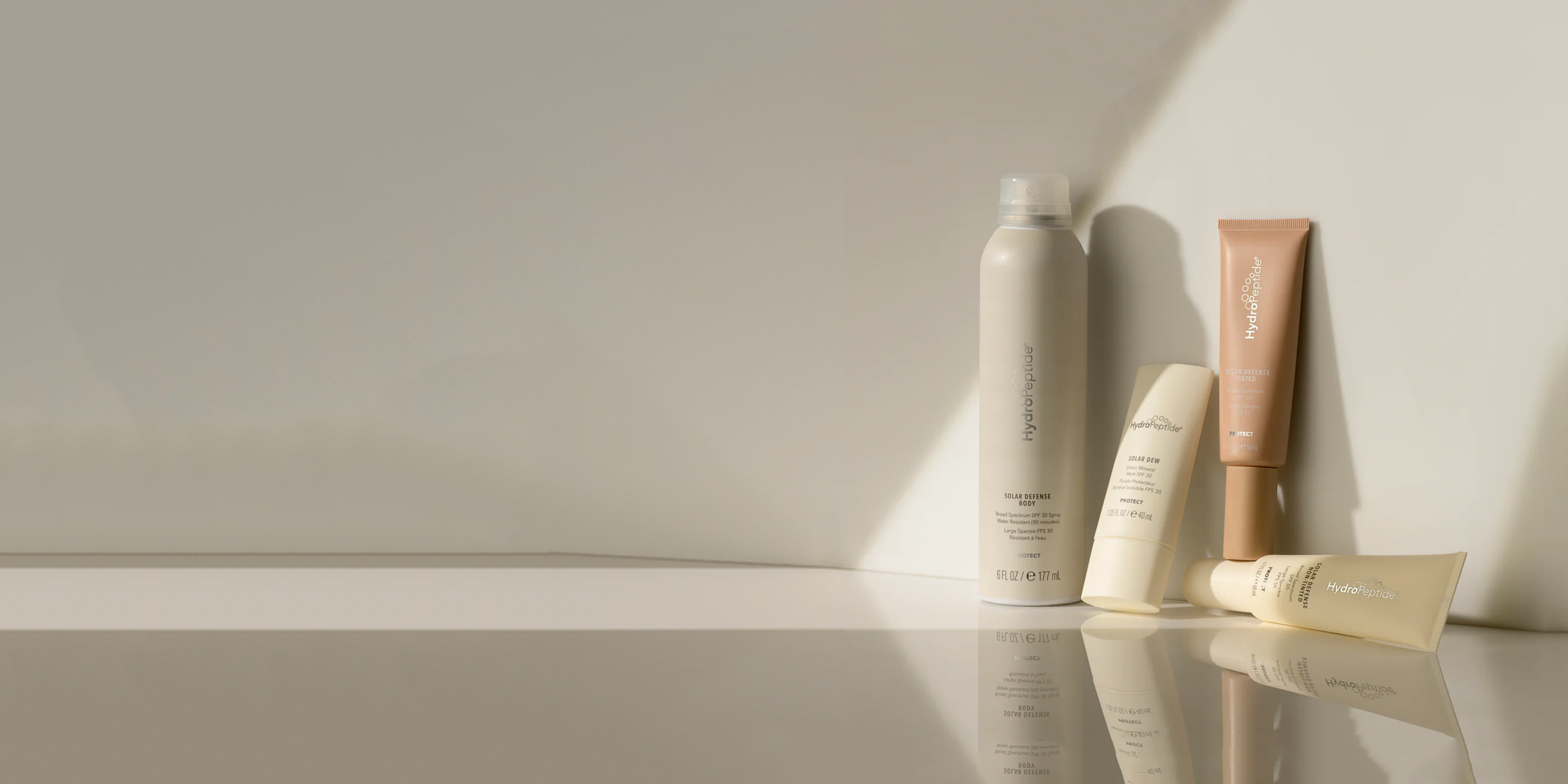 Skincare products from Hydropeptide in minimalist beige packaging are arranged on a reflective surface. The composition includes a spray can and various tubes, set against a plain, light coloured background with soft shadows.