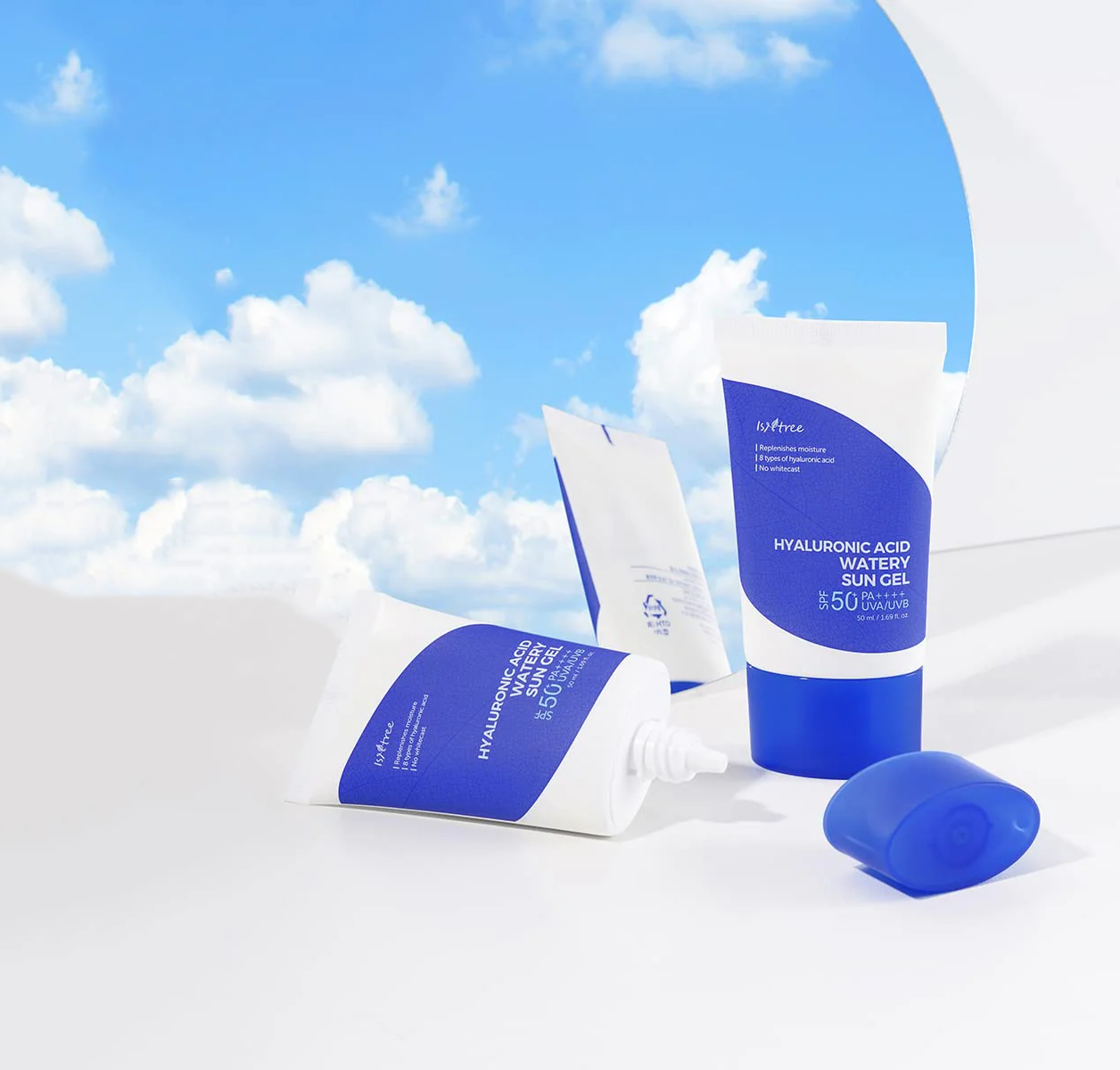 Skincare products from Isntree arranged on a white surface against a backdrop of a blue sky with fluffy clouds. Tubes and packets, primarily in white and blue, are labeled Hyaluronic Acid Watery Sun Gel with SPF 50.