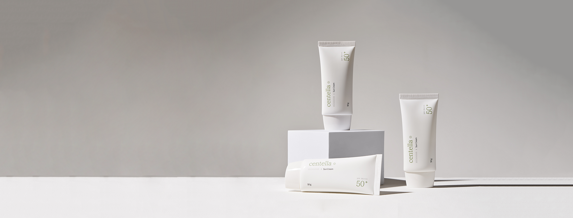 Three tubes of skincare products from Mixsoon labeled centella are displayed against a neutral background. One tube is elevated on a small white pedestal, while the other two are placed directly on the surface.