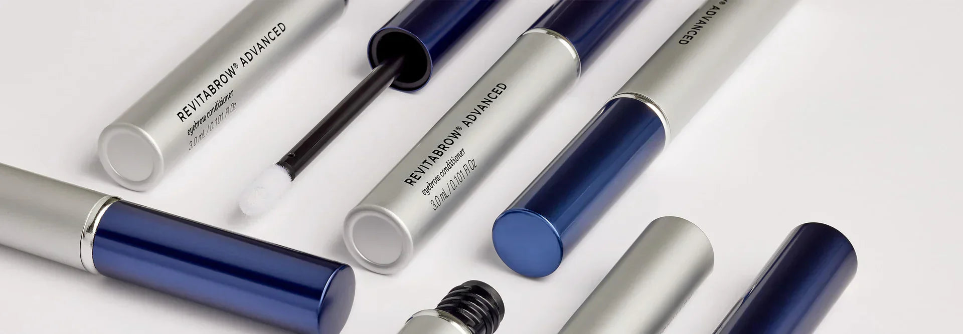 Assortment of silver and blue cosmetic tubes from Revitalash labeled RevitaBrow Advanced on a light background, with some tubes open to reveal applicators.