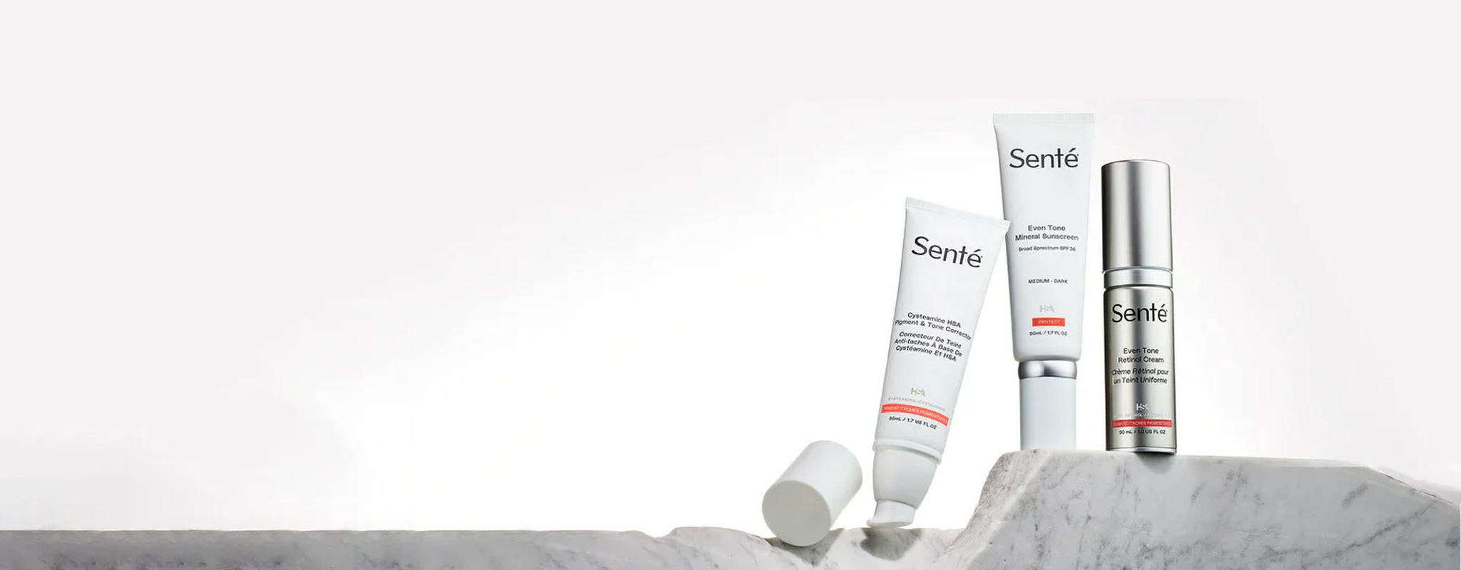 A set of Senté skincare products displayed on a marble surface. Three items include two tubes and one bottle, featuring minimalist white packaging with text and a hint of orange. The background is a light grey, creating a clean, modern look.