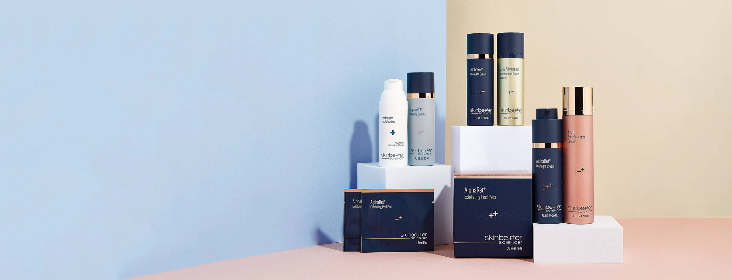 A selection of skincare products from Skinbetter Science displayed on geometric stands against a two-tone background. The arrangement includes bottles and boxes in navy and white packaging with gold accents.