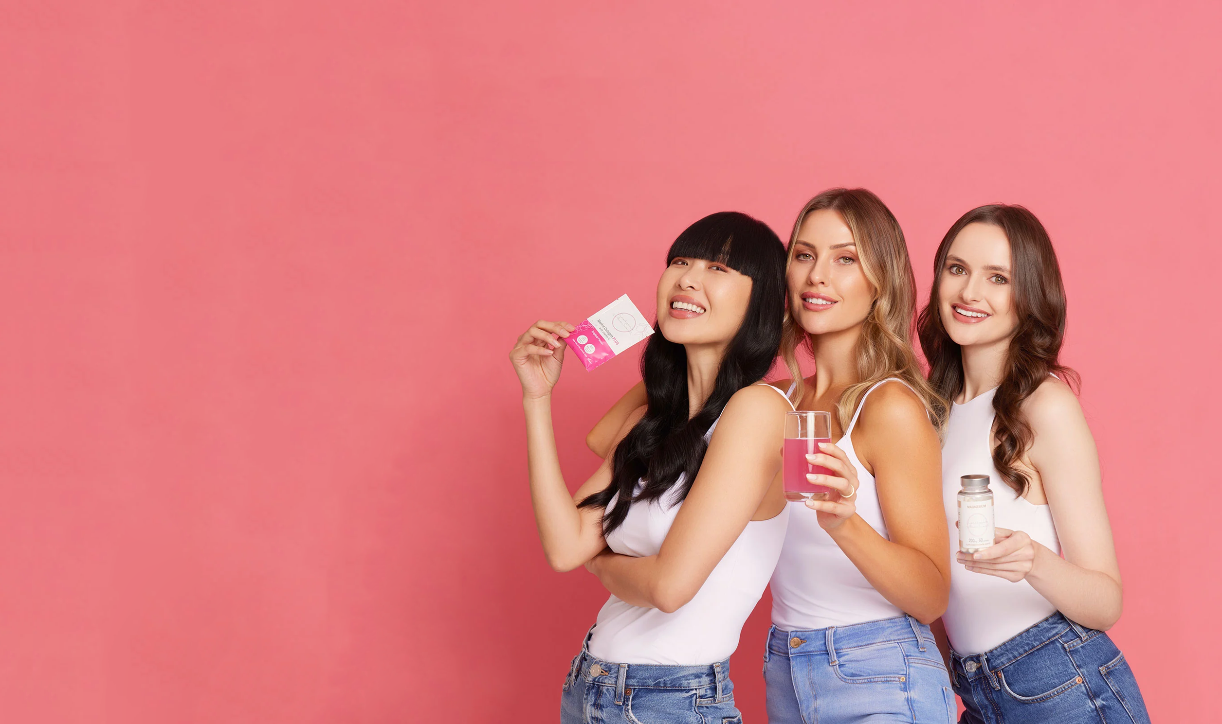 Three women in white tops and jeans stand against a pink background, smiling. One holds a packet, another has a glass with a pink drink, and the third has a jar. They appear joyful and relaxed holding Supplements Made Simple products.