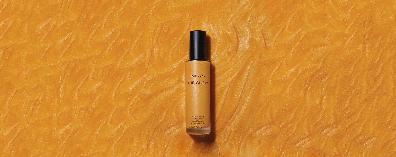 A bottle of The Glow illuminating liquid from Tan-Luxe is centered on a textured, vibrant orange background. The bottle is sleek with a black cap, and its warm, golden hue complements the backdrop.