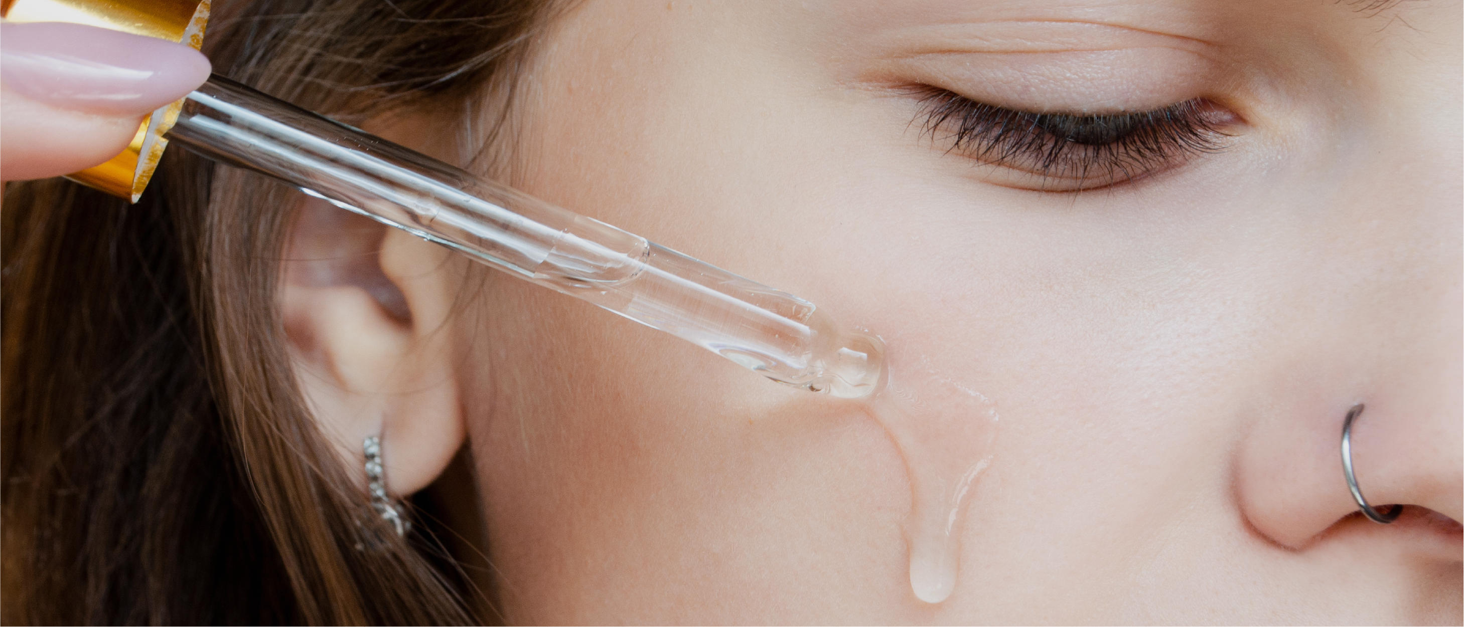 Close-up of a young face with nose-ring holding a serum dropper emitting clear gel on the cheek.