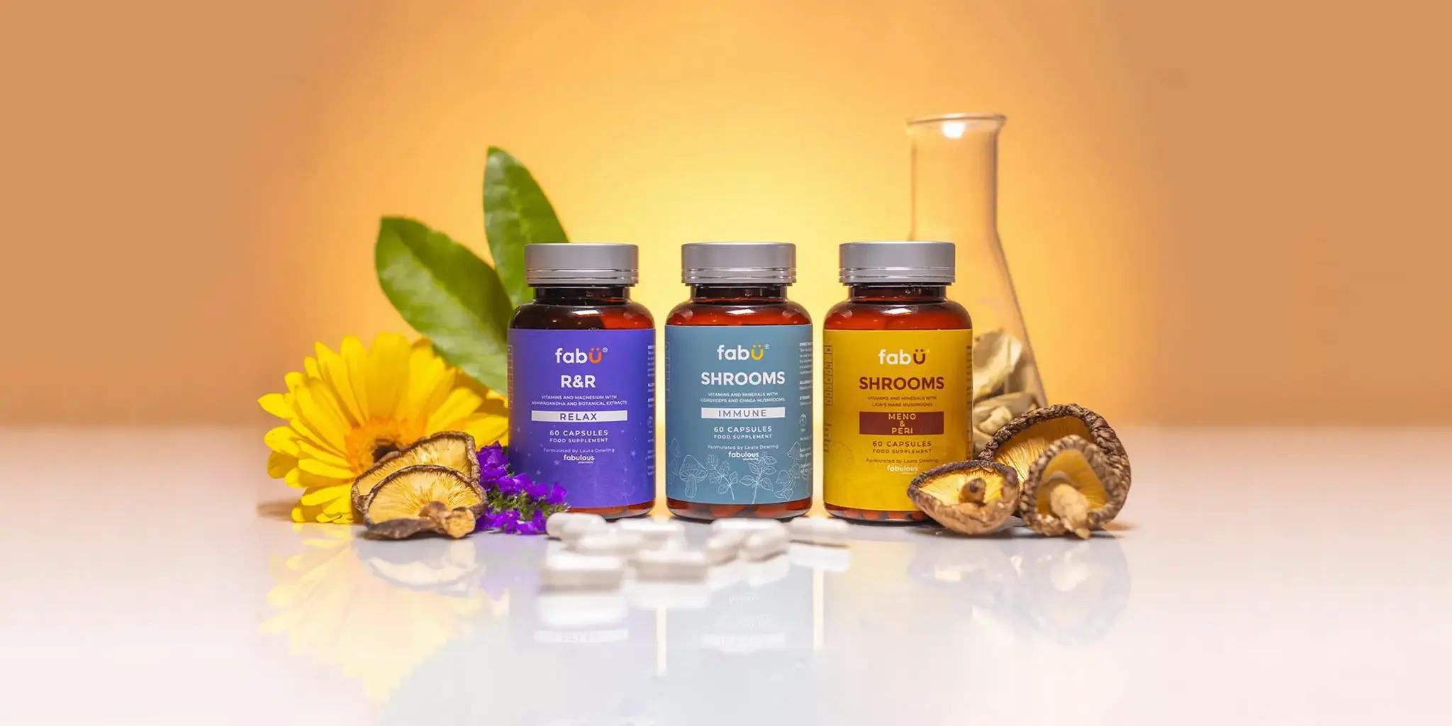 Three bottles of dietary supplements labeled fabÜ are on display with a sunflower, purple flowers, leaves, mushrooms, and white capsules around them. The background is a warm, gradient orange.