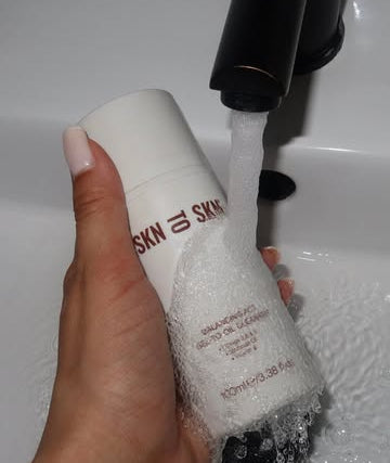 A hand holds a white bottle labeled SKN TO SKN CLEANSER 
under a black faucet with running water, with overlaid text reading 'THE 5-MINUTE EVENING ROUTINE'.