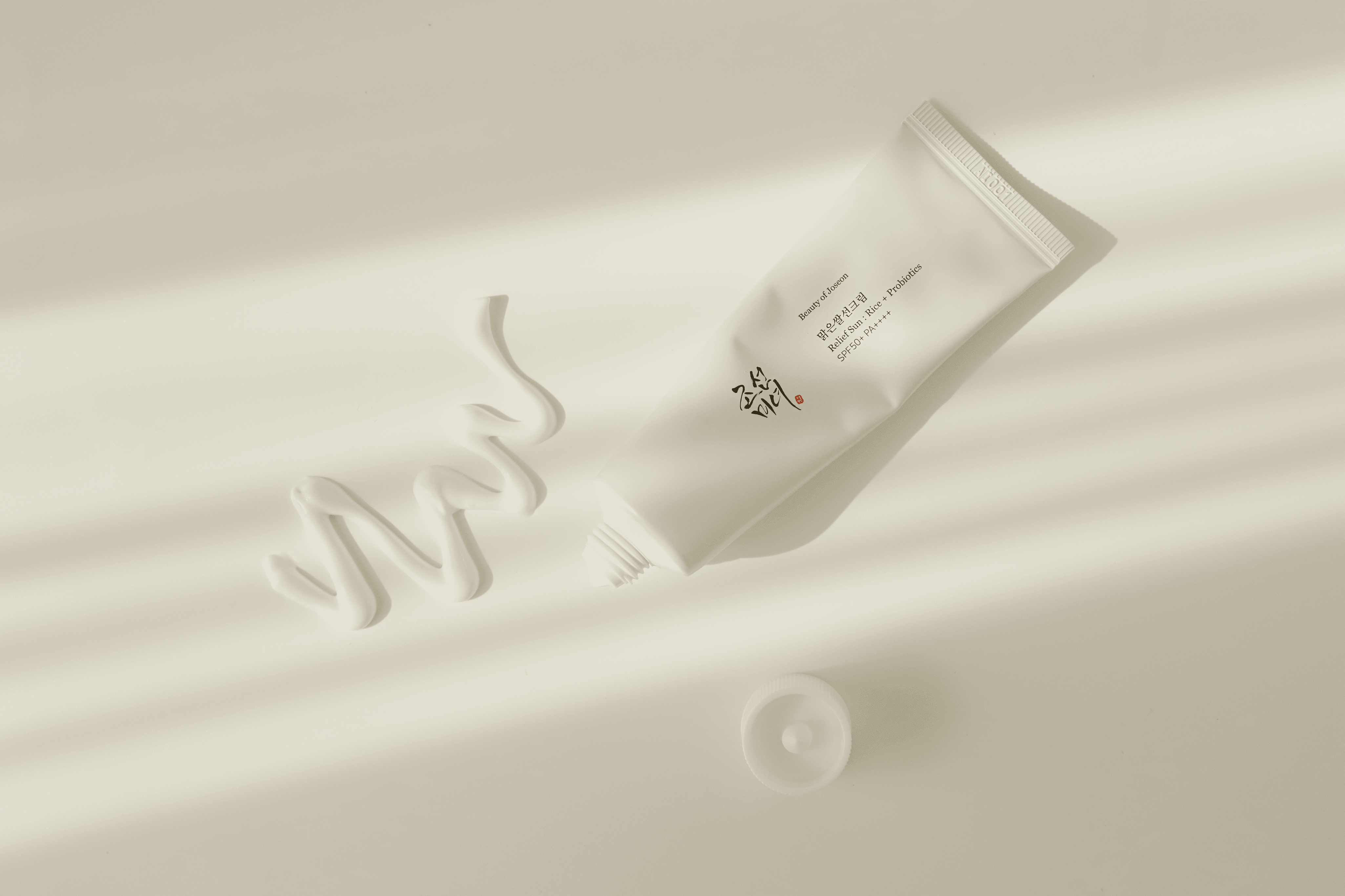 A white tube of cream from Beauty of Joseon with its cap off lies on a sunlit surface. Cream is artistically squeezed out in a wavy line next to the tube.