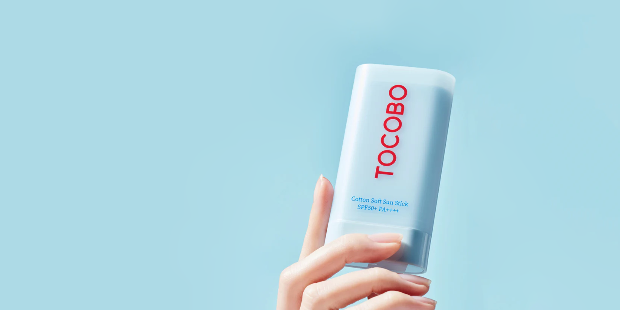 A hand holding a light blue sun stick container with the brand name TOCOBO in red. The label reads Cotton Soft Sun Stick SPF50+ PA+++. The background is a solid light blue.